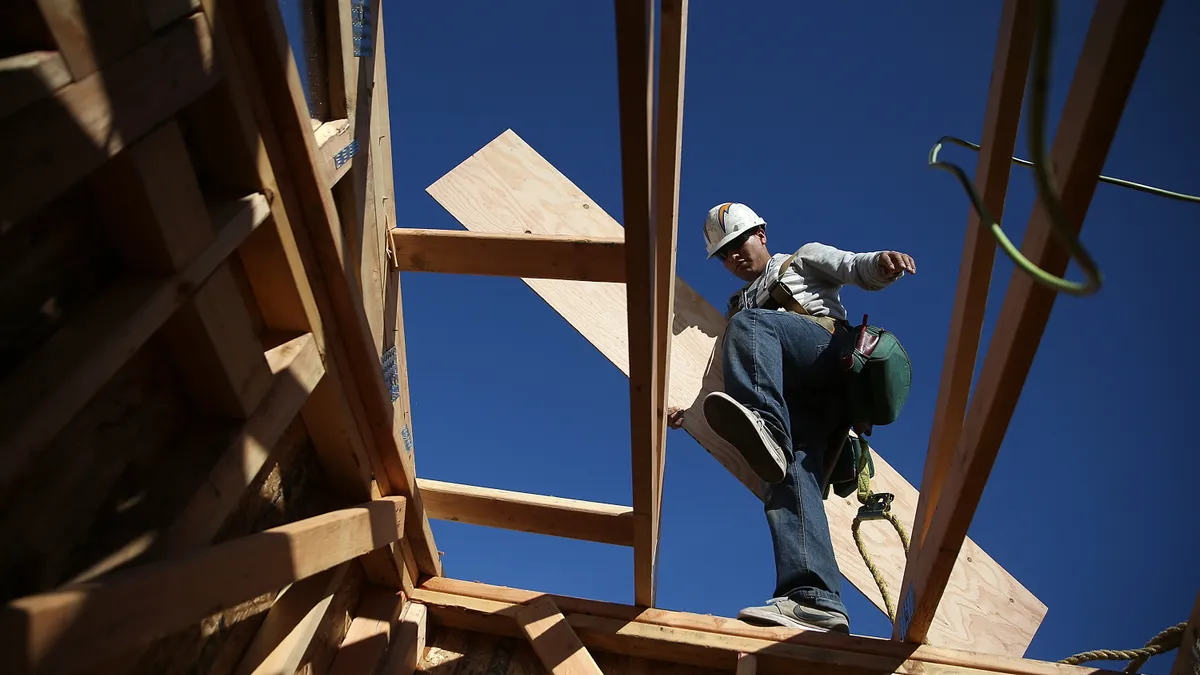 A construction worker as seen from below, stepping over a gap while carrying a piece of lumber.