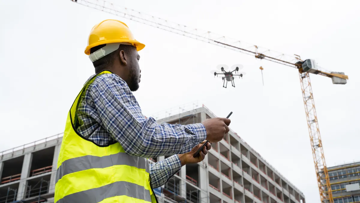 A person in a collared shirt and safety equipment looks up at a drone flying above them.