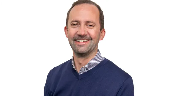 A smiling white man in a blue v-neck sweater in front of a stark white background.