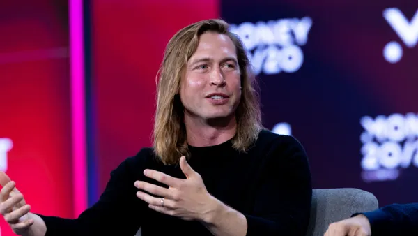 A person seated on a stage gestures while speaking.