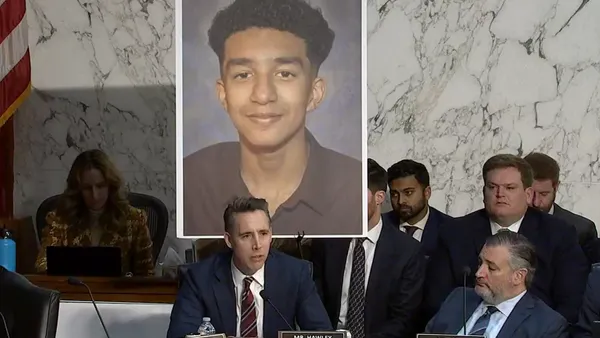 A senator speaks during a committee hearing with a sign of a teenager's headshot behind him.