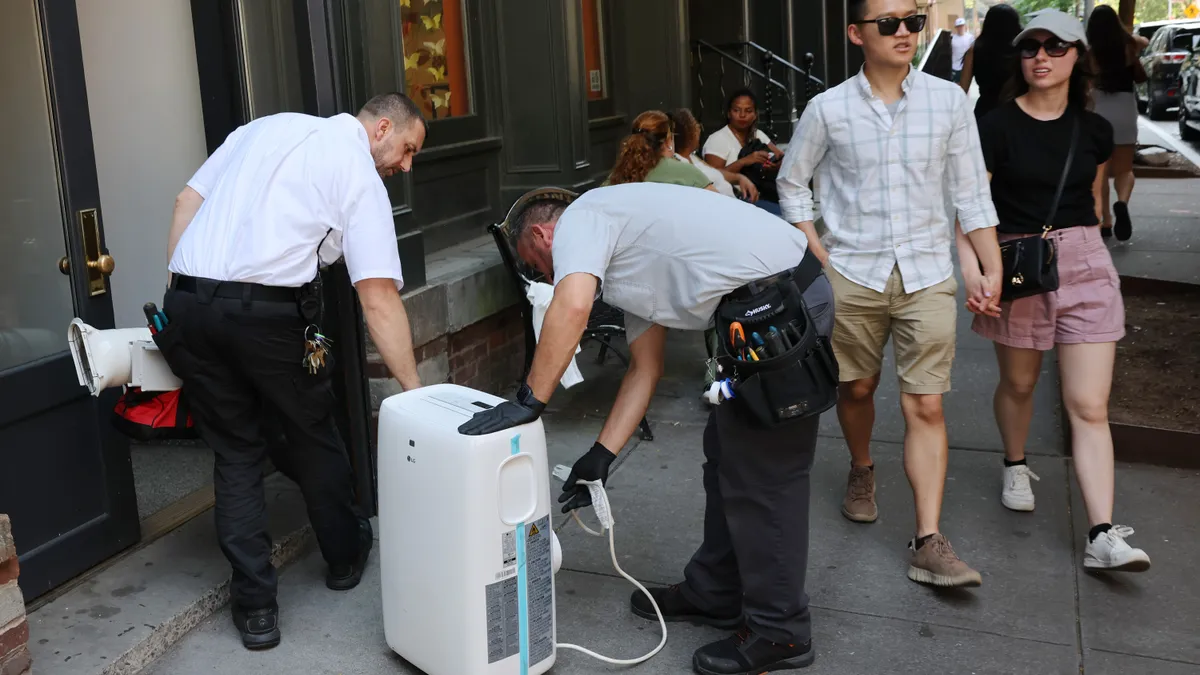 Two people carry equipment into a building on a busy city sidewalk.