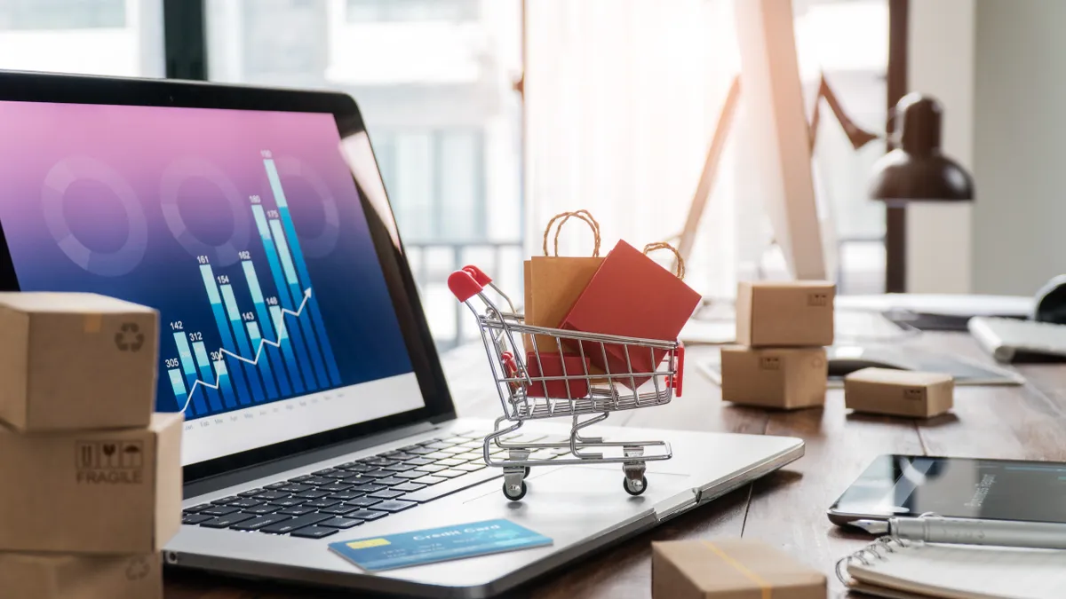 Shopping bags incart and credit card on laptop with paper boxes on table and sales data economic growth graph on screen.