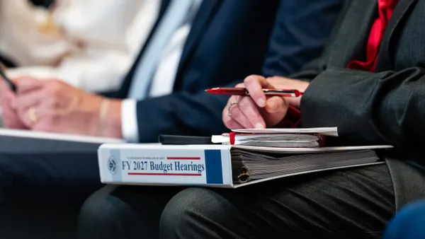 A white binder with "FY27 Budget Hearings" written on the spine in the lap of a person wearing a blue suit.
