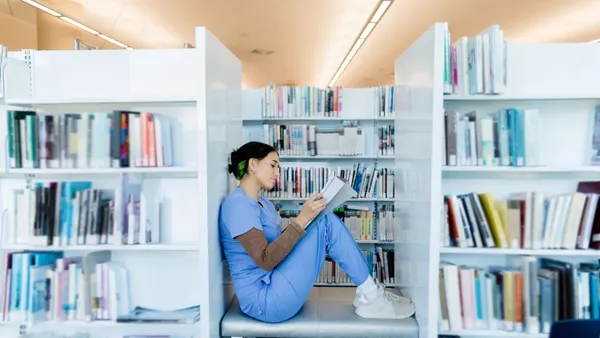 A person in scrubs sits between two bookshelves, reading a book.