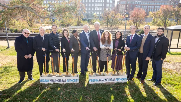 12 men and women holding shovels on a grassy area with railroad tracks and buildings in the background.
