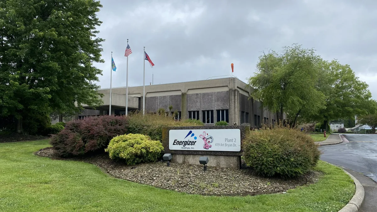 A white Energizer sign with the pink Energizer bunny and a brown-gray building behind it surrounded by green landscape, and a parking lot on a cloudy day.