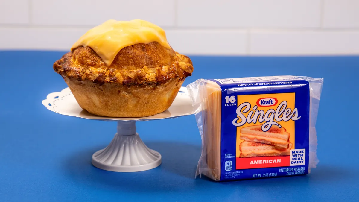 A single-serve apple pie with a slice of Kraft Singles melted on top sits on a doily on a small white pie stand. A package of Kraft Singles sits to the right on a blue table.