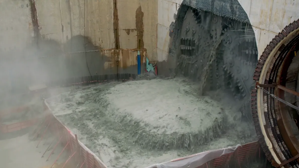 A massive flat circular machine bores through a tall concrete wall while grey water gushes underneath.