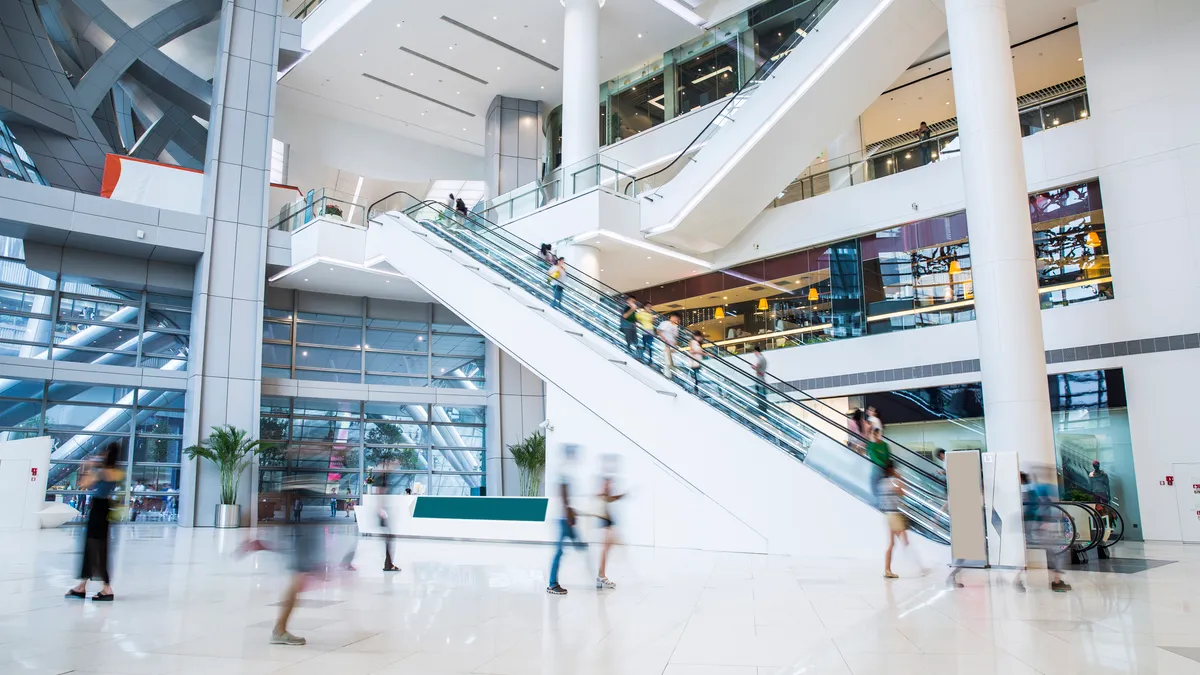 People blurred walking through a shopping mall forum with escalators in the background.