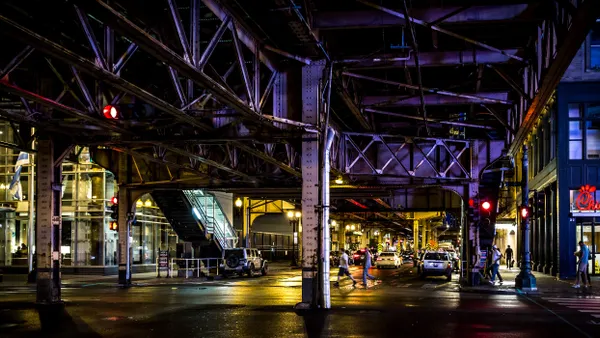 People cross a street underneath elevated train tracks at night.
