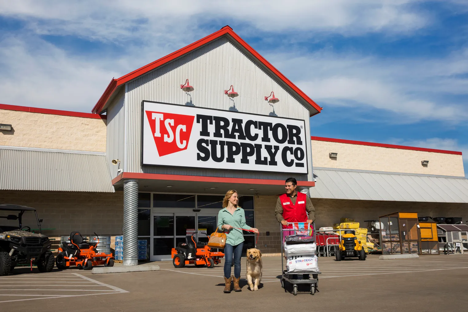 Two smiling people, one with a dog on a leash and the other with a shopping cart, walk away from a store entrance.