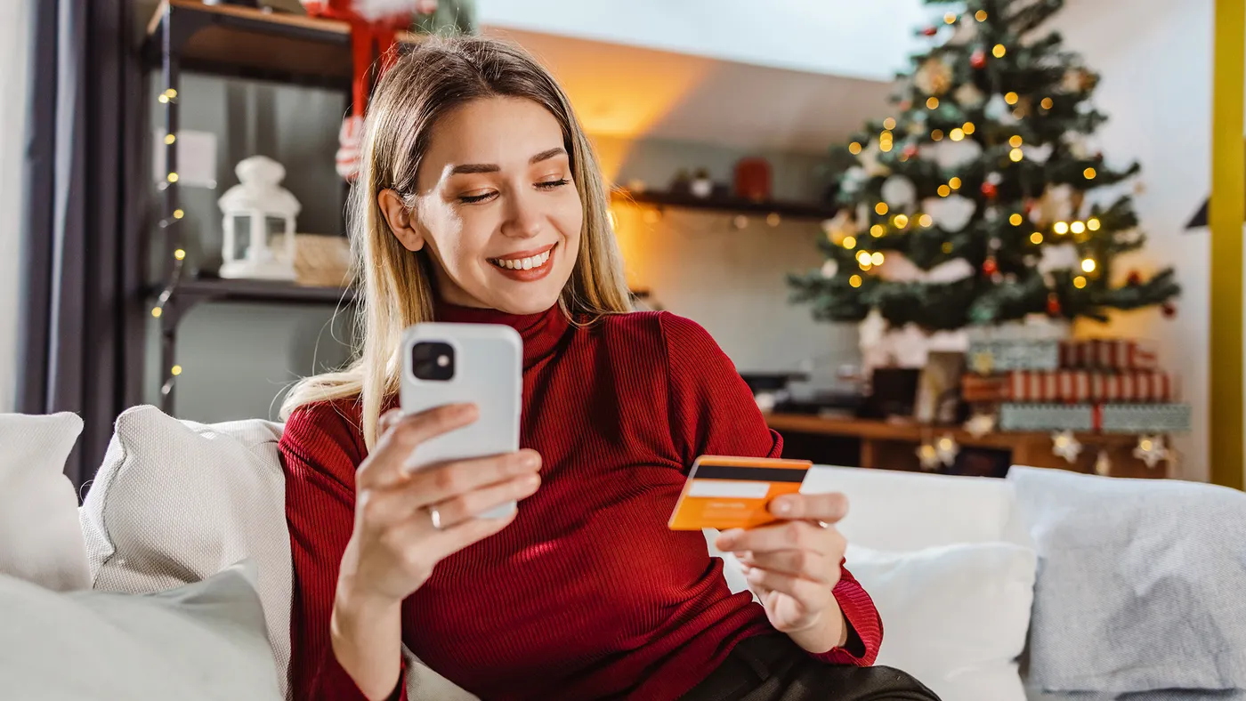 Person smiling while shopping online with a smartphone and credit card in a festive, holiday-decorated living room.