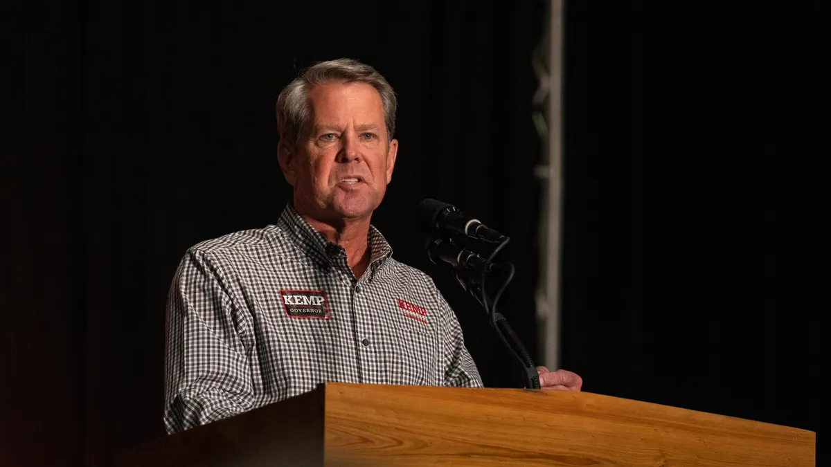 Gov. Brian Kemp stands behind a podium at an election event wearing a checkered shirt.