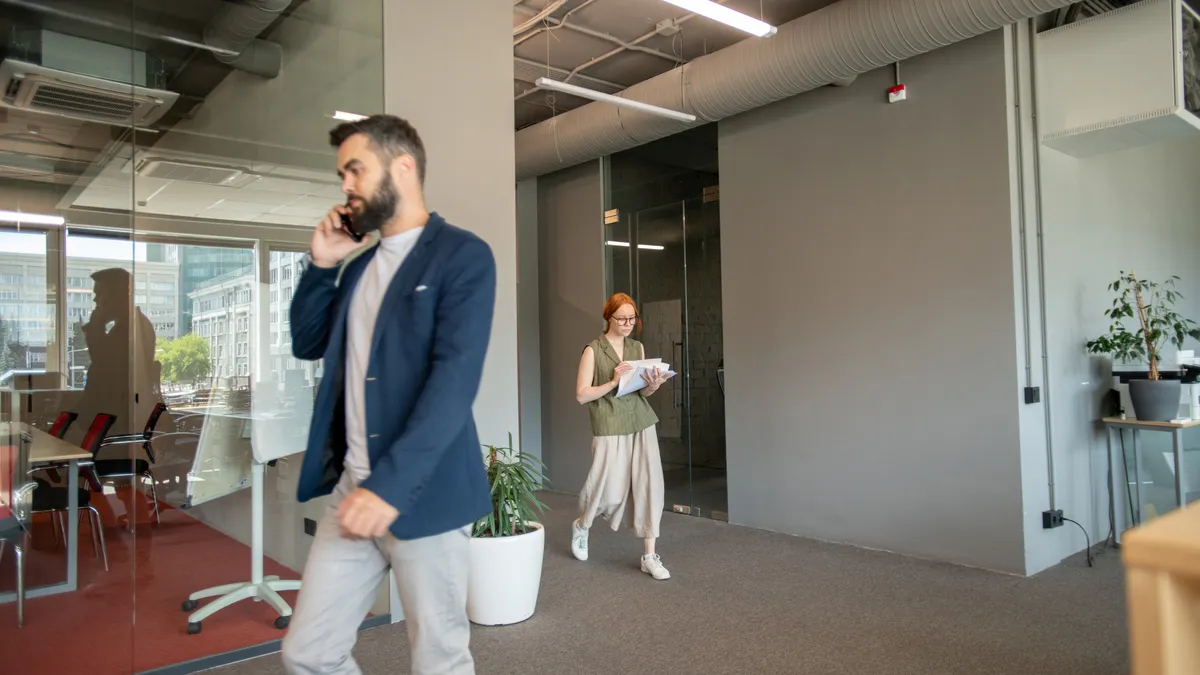 Young businessman in smart casualwear talking on mobile phone in office while his colleague with papers moving towards her workplace