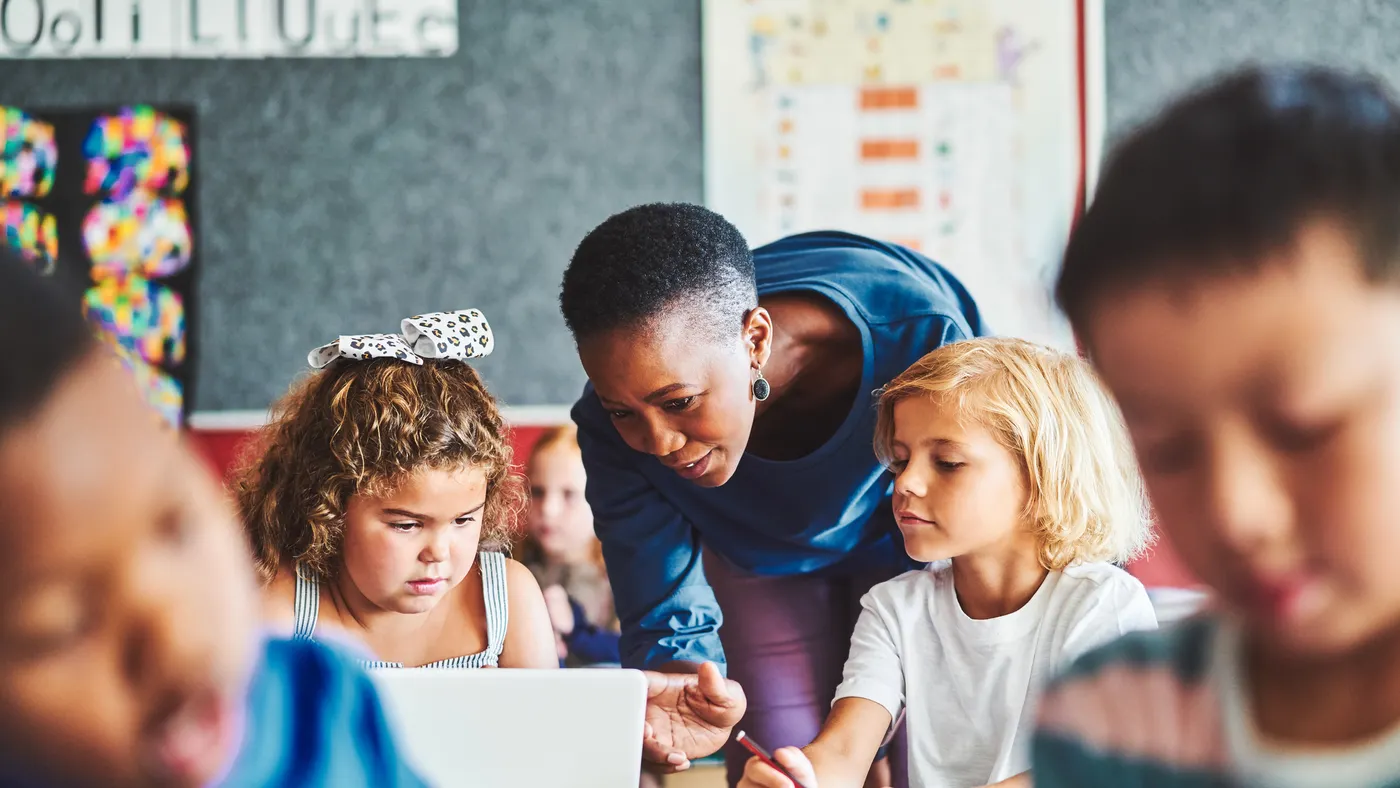 Several students are sitting at desks in a classroom. A teacher who is standing is leaning down to help a student on a laptop.