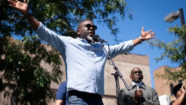 A man stands on a podium at a microphone raising his arms in the air.