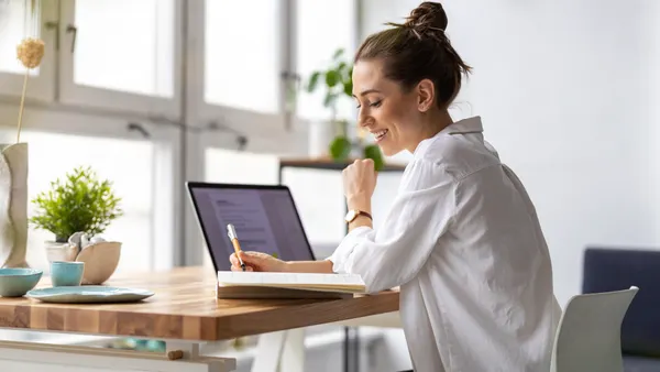 A person at a desk and writing in a notebook with an open laptop next to them