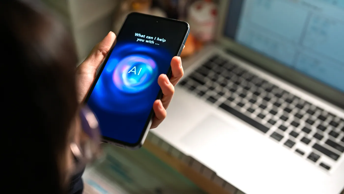 A person uses an AI chatbot on a smartphone while sitting in front of a computer