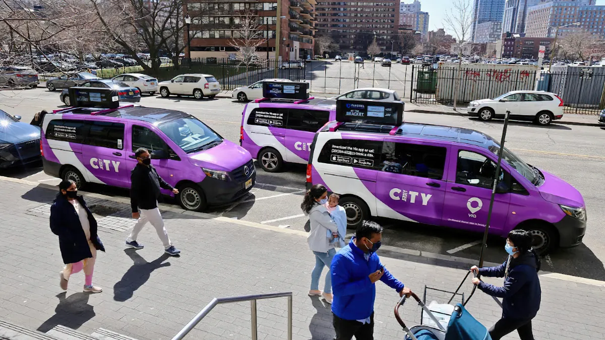 Several people wearing masks are walking along a sidewalk with three purple and white minivan parked at the curb.