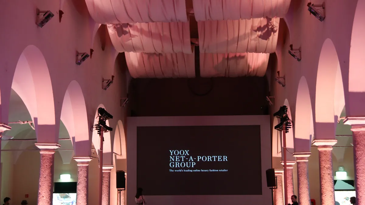 Draped pink curtains hang from the ceiling and pink walls with large arched columns frame a screen reading Yoox Net a Porter Group.