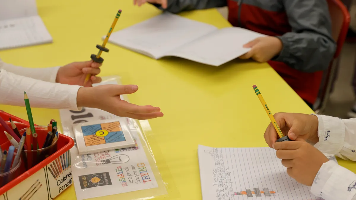 A young student's hand reaches across a desk toward another student's hand.