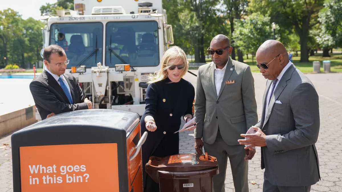 Four city officials standing around an organics recycling bin, with a white sanitation truck in the background