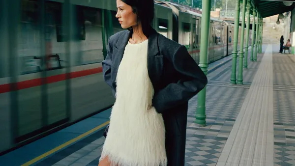 A model stands on a train platform wearing a white Anthropologie dress and oversized gray coat