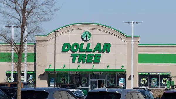 A Dollar Tree store with a large green and white sign above the entrance, located in a shopping center. The building has a beige exterior with green trim and large windows displaying promotional signs. Several parked cars are visible in the foreground, and a leafless tree stands on the left side of the image under a clear blue sky.