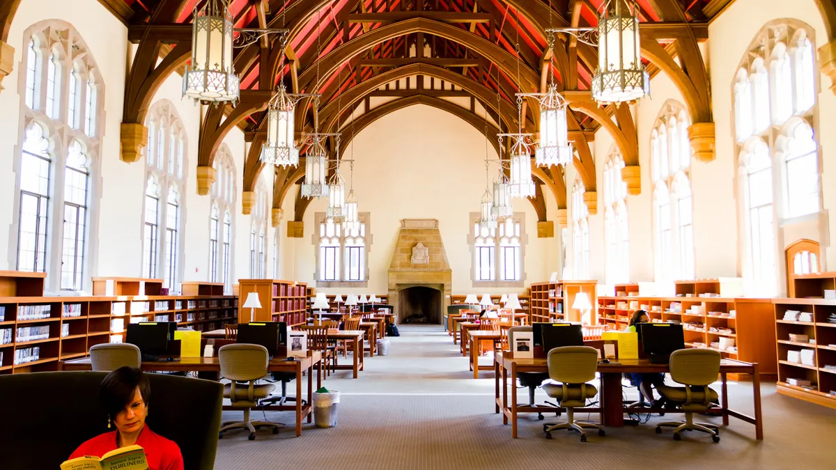 A person reads a book in a room with tall windows, a high-arching ceiling, a fireplace, tables, and shelves of books.