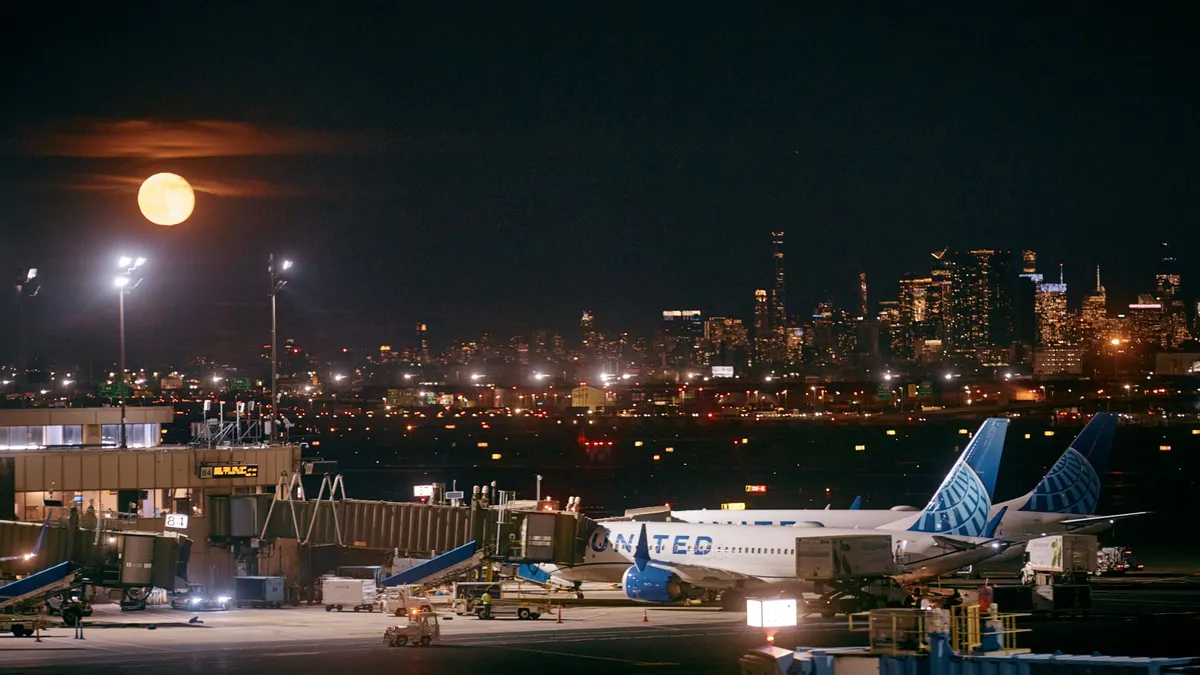 A plane is loaded at an airport with the New York City skyline and the moon in the background.