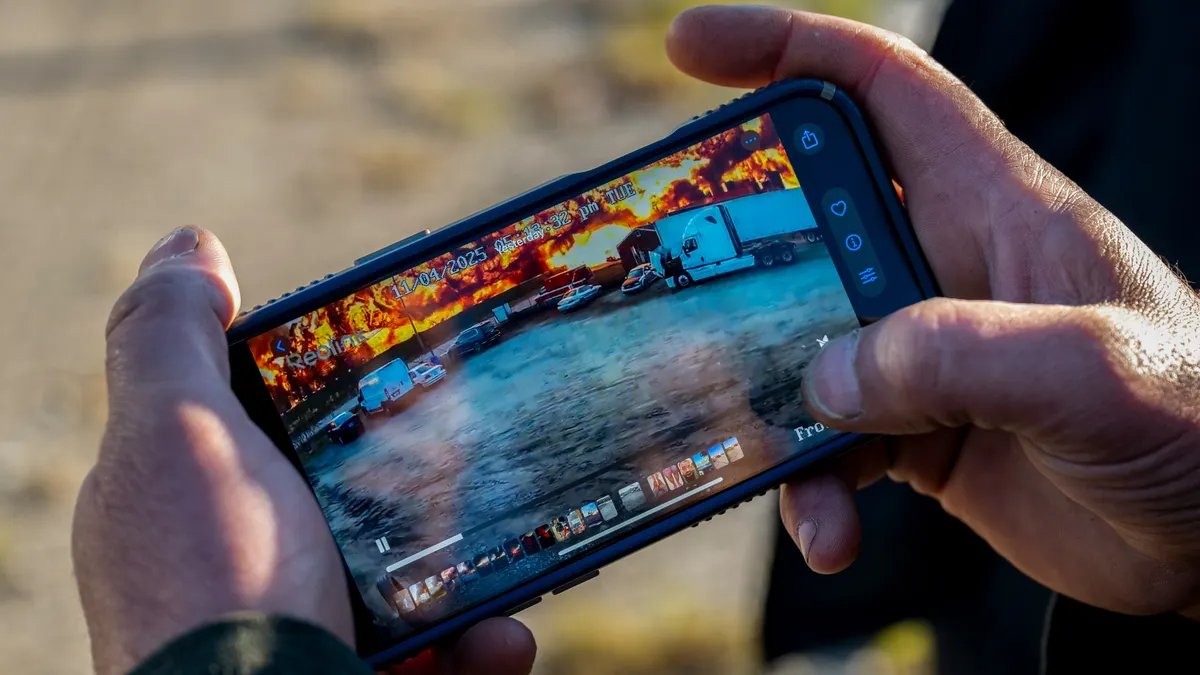Hands hold a phone showing a video with vehicles in the foreground and a blazing fire in the background.