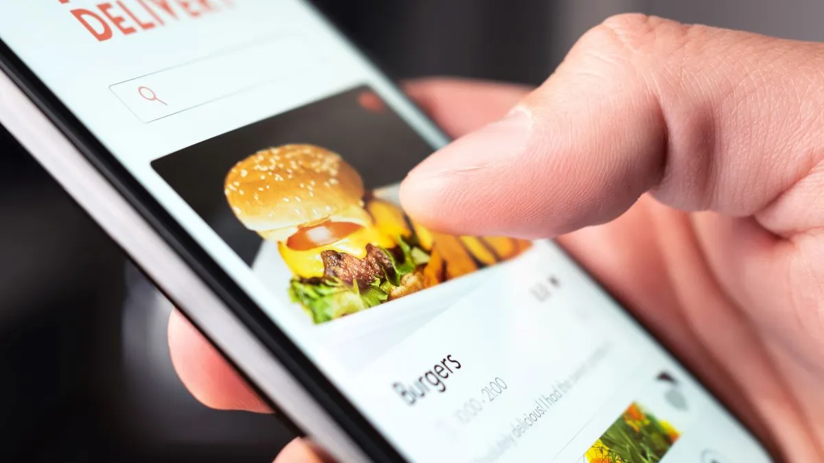 A thumb hovers over a smart phone with a photo of a cheeseburger and the words food delivery on top.