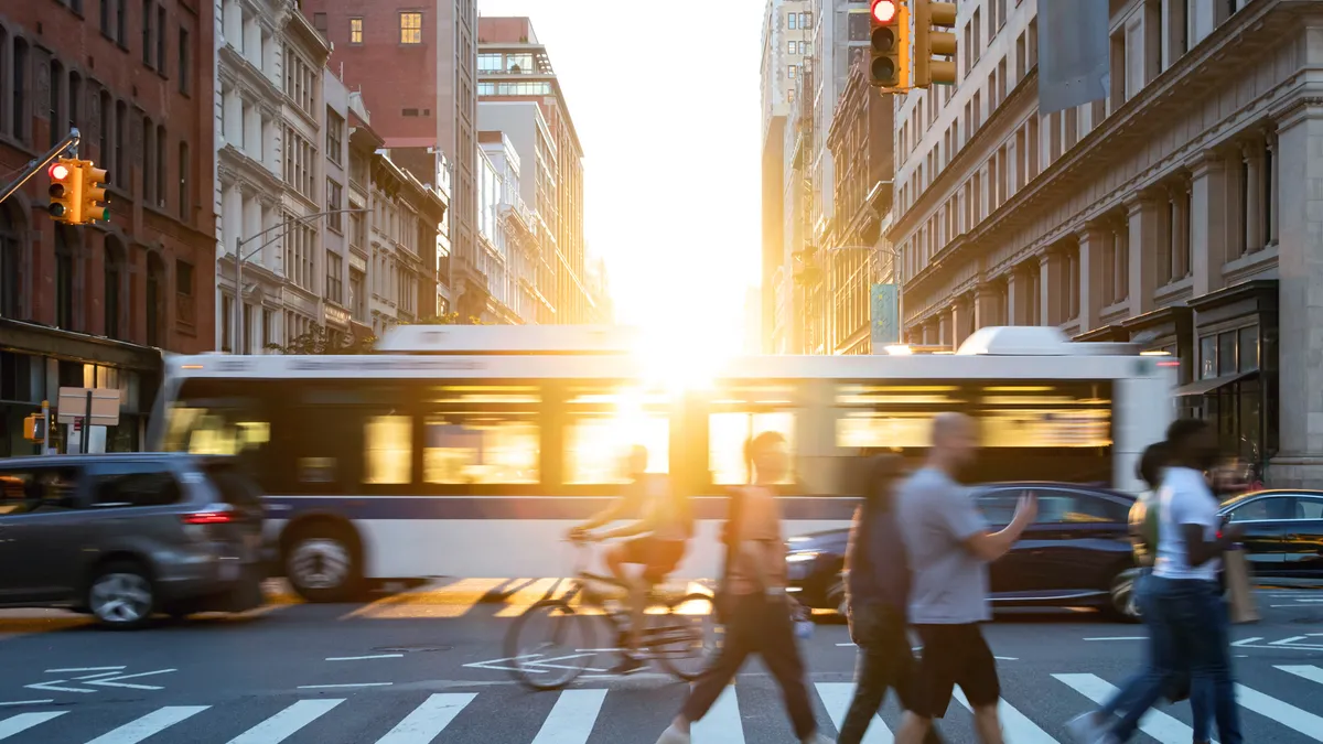 Pedestrians and a cyclist cross a crosswalk at a busy city intersection with a burst of sunlight shining through.