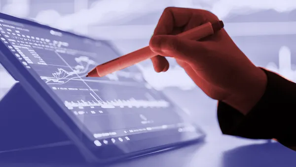 A person holds a stylus against a tablet standing upright as a display. The tablet shows graphs and charts indicating financial planning.