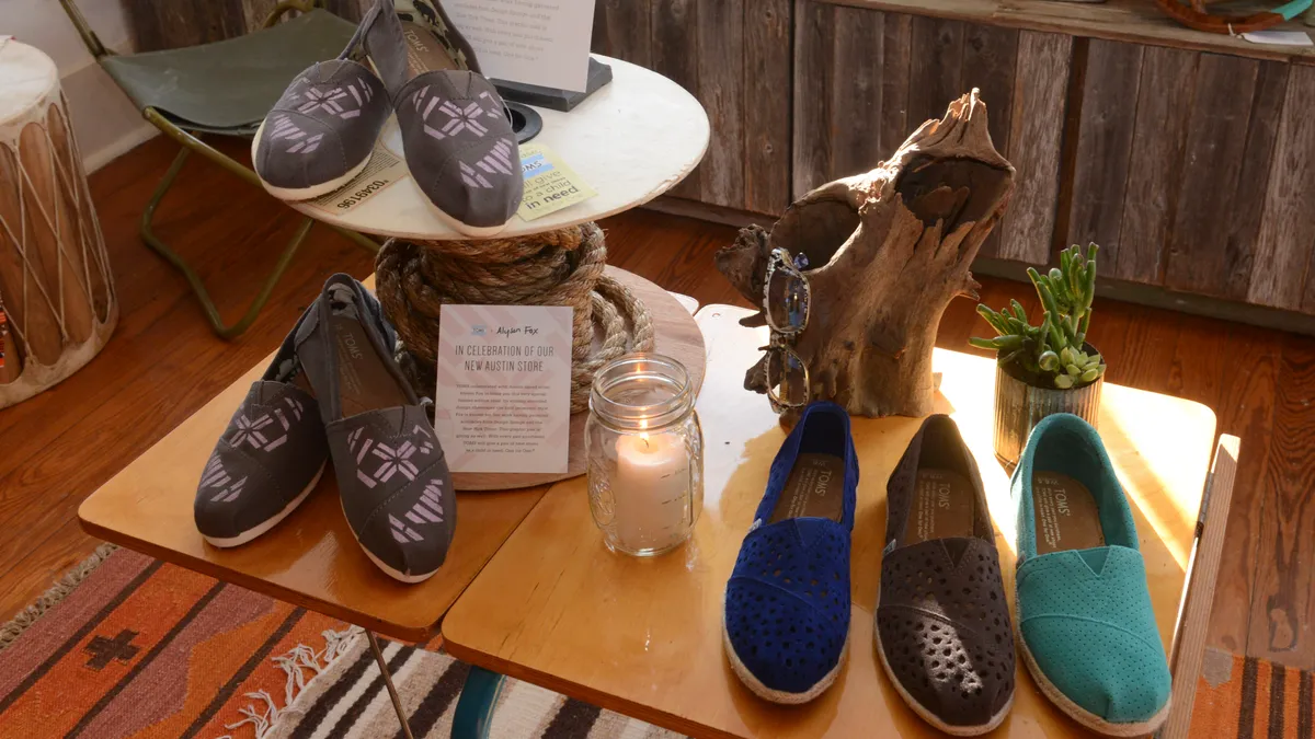 A store display for Toms. It features several Toms slip-in styles, including a grey with white checked pattern. Other shoes on display are dark blue, brown and teal.