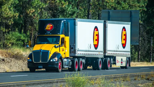 An Estes Express Lines semi-truck pulling double trailers on a wooded section of freeway.
