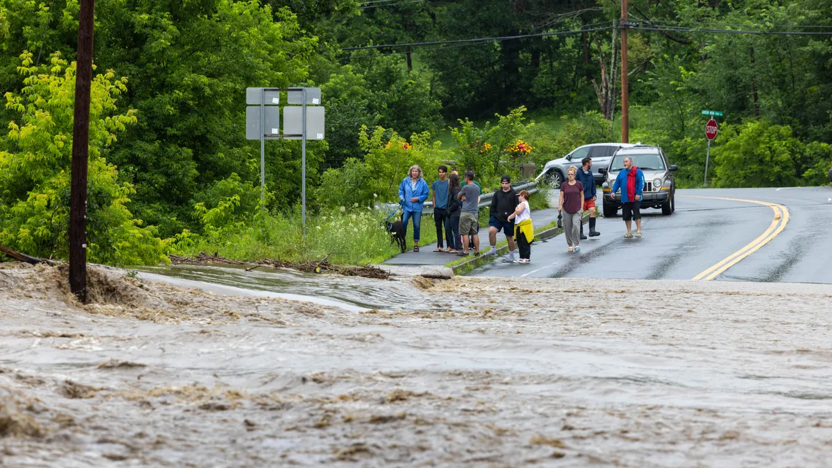 A group of people and cars stand at the edge of a flooded road.