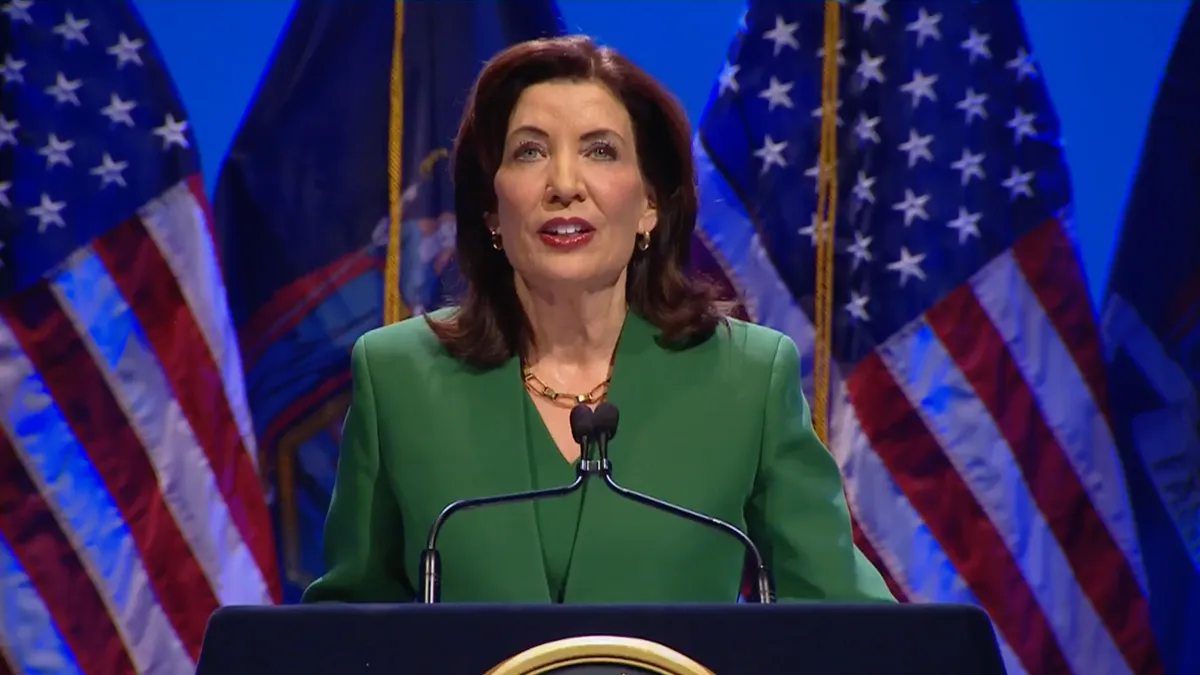 A brunette woman in a green suit speaks at a podium in front of several U.S. flags.