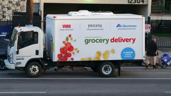 A medium-sized white delivery truck is parked along a street in front of a beige building. The truck is wrapped with colorful images of tomatoes and citrus fruits and branded with logos for Vons, Pavilions, and Albertsons. A man is seen with a dolly unloading blue crates.