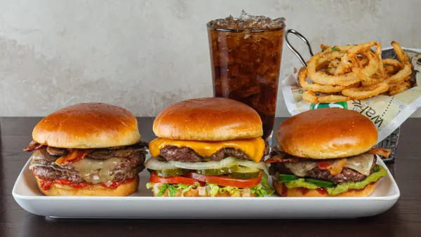 An image of three burgers on a white place in front of a basket of curly fries and a glass of brown soda.