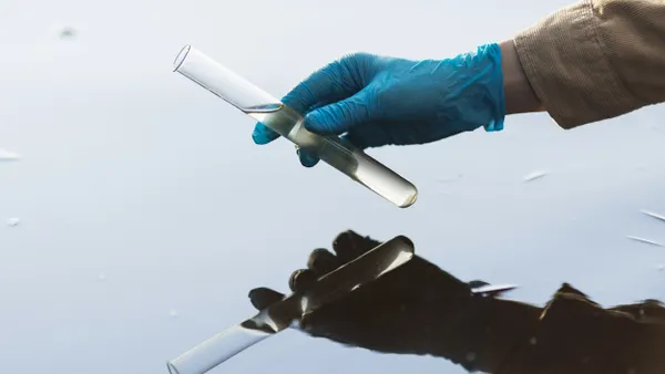 A gloved hand holds a test tube with yellowish water reflected in a body of still water.