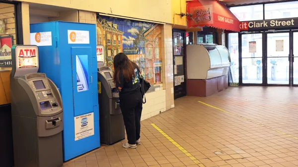 A person with their back to the camera conducts a transaction at an ATM in a subway station. A bitcoin ATM and another ATM sit next to the first machine.