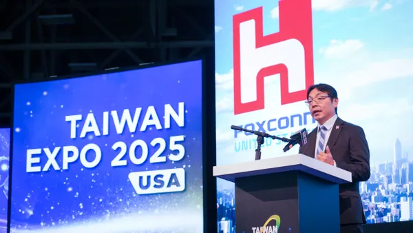 A dark-haired person with glasses speaking into a mic at a podium with the Taiwan Expo USA on the left of him.