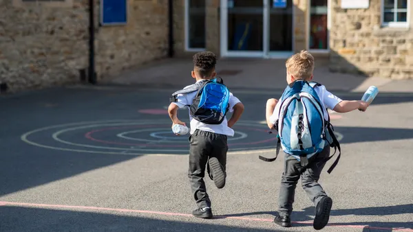 Two students run in a schoolyard with their backpacks on