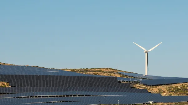 A large solar panel farm spread across a hillside with a single wind turbine in the background under a clear blue sky.