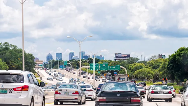 Passenger cars are several lanes of highway with a skyline of city buildings during a sunny day in the background.
