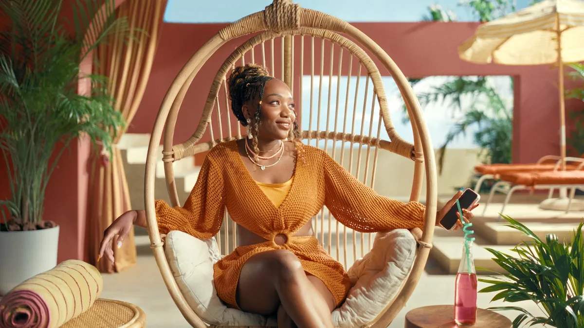 A woman wearing an orange dress sits on a chair against a tropical backdrop.