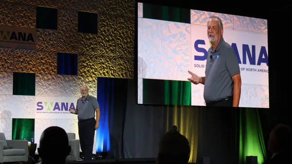 A man wearing a polo stands on a stage with a live feed projected behind him. The stage backdrop is decorated with the SWANA logo.