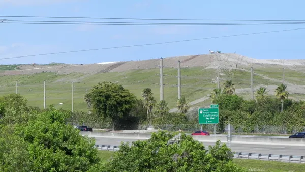 A landfill covered in gras rises above a highway. A sign on the road notes an exit for the South Sawgrass Expressway toward Coral Springs and Key West.